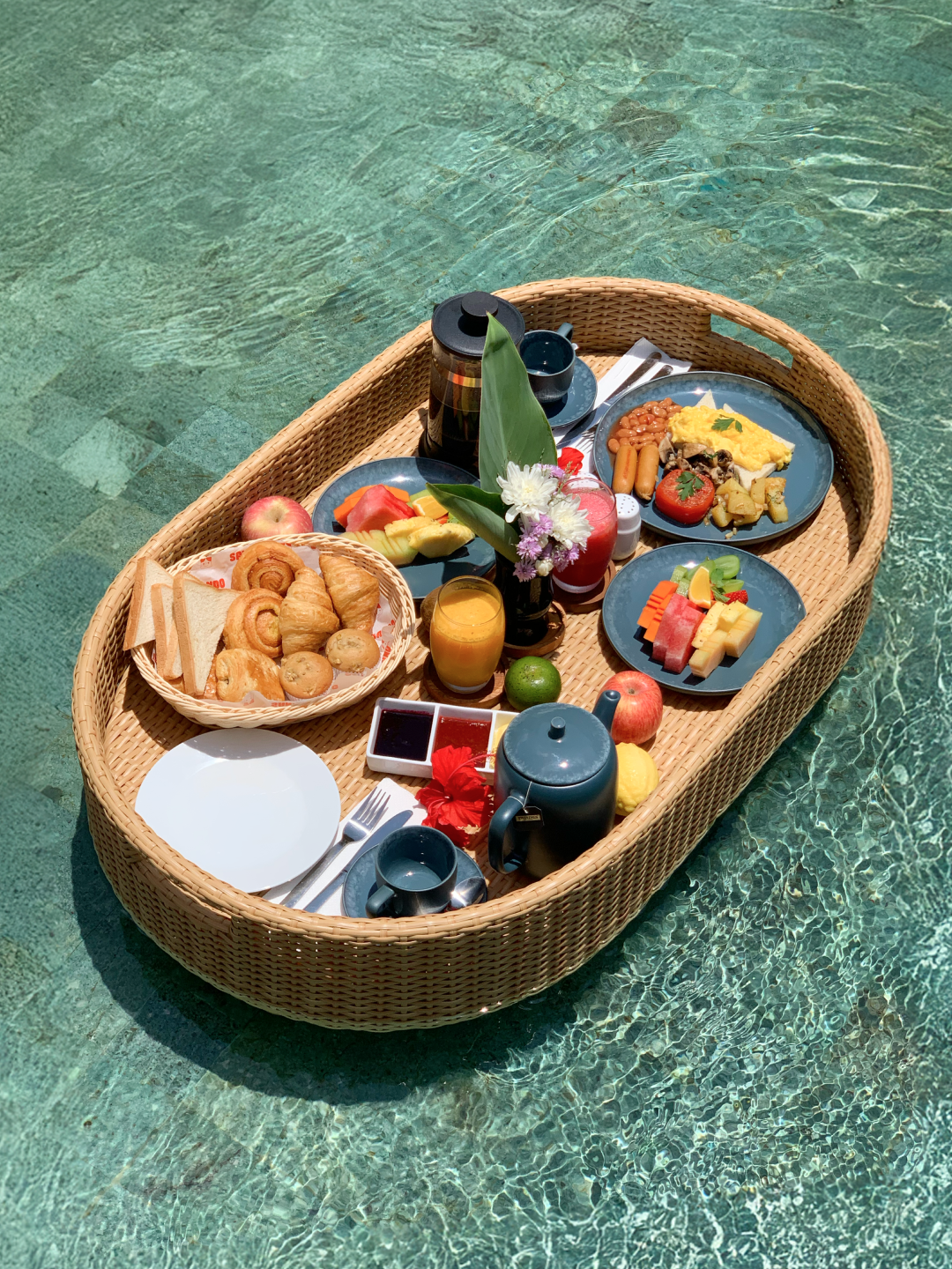 Floating breakfast tray with fruit, pastries, coffee, and drinks in clear turquoise water