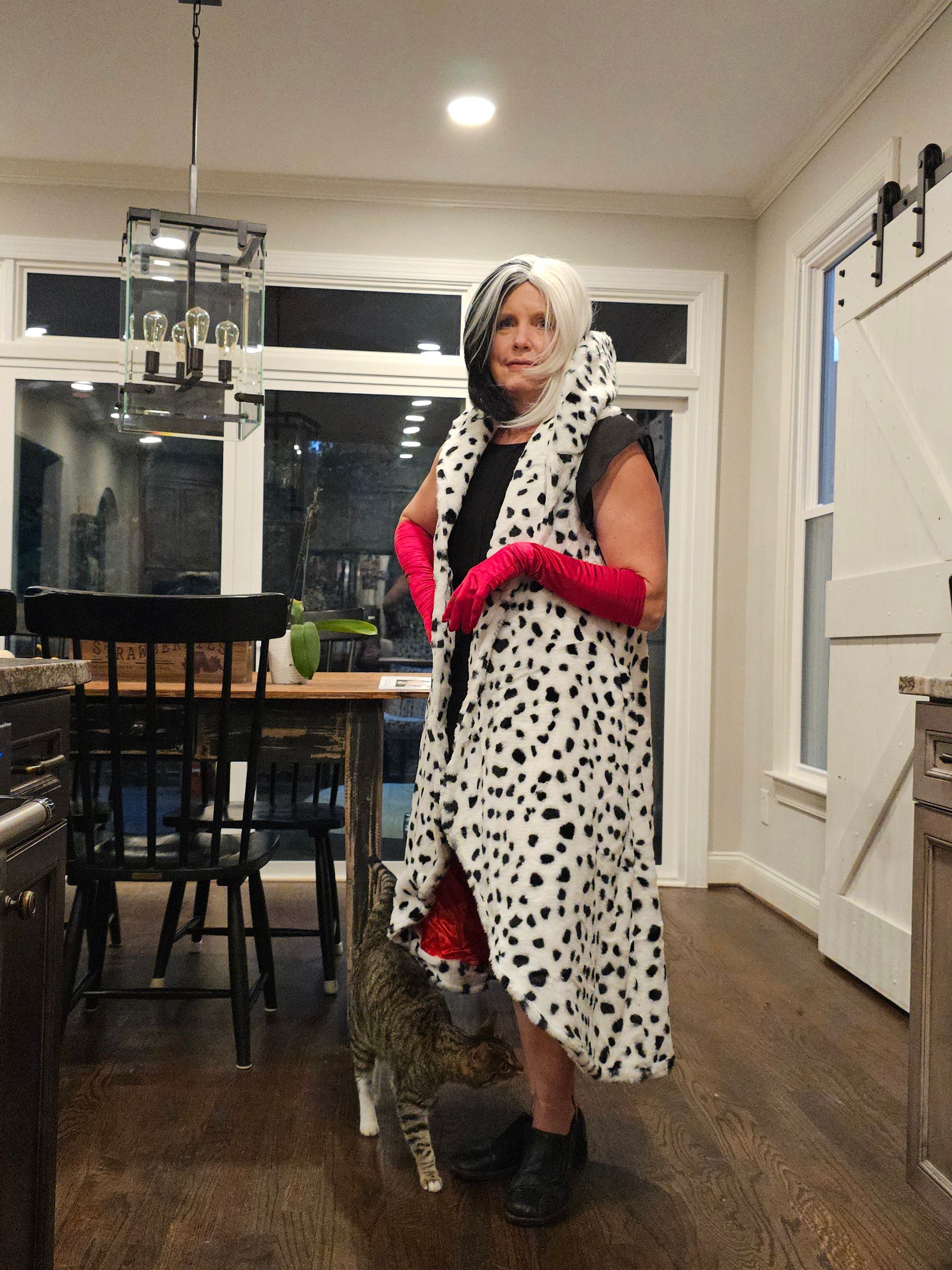Person standing indoors in a spotted white outfit with bright pink gloves beside a dining table
