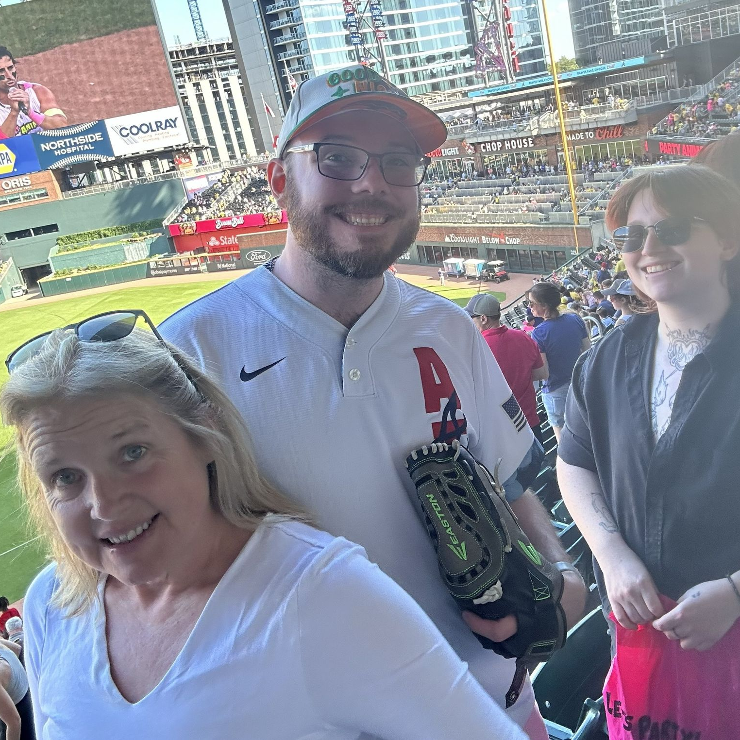 Three fans smiling at a baseball stadium, with the field and stands in the background.