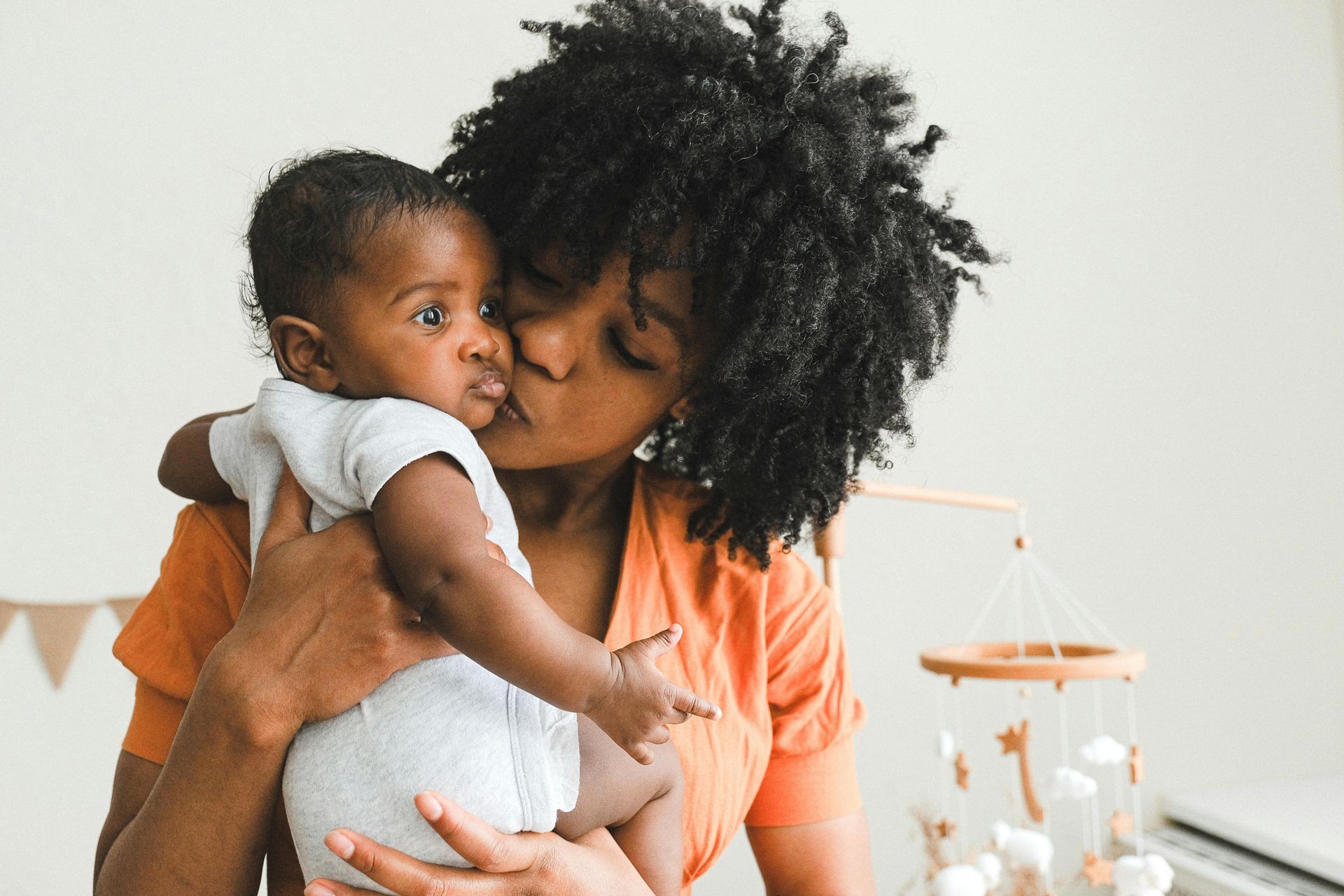 Woman holding and kissing a baby in a white room, with an orange shirt and a small lamp in the background