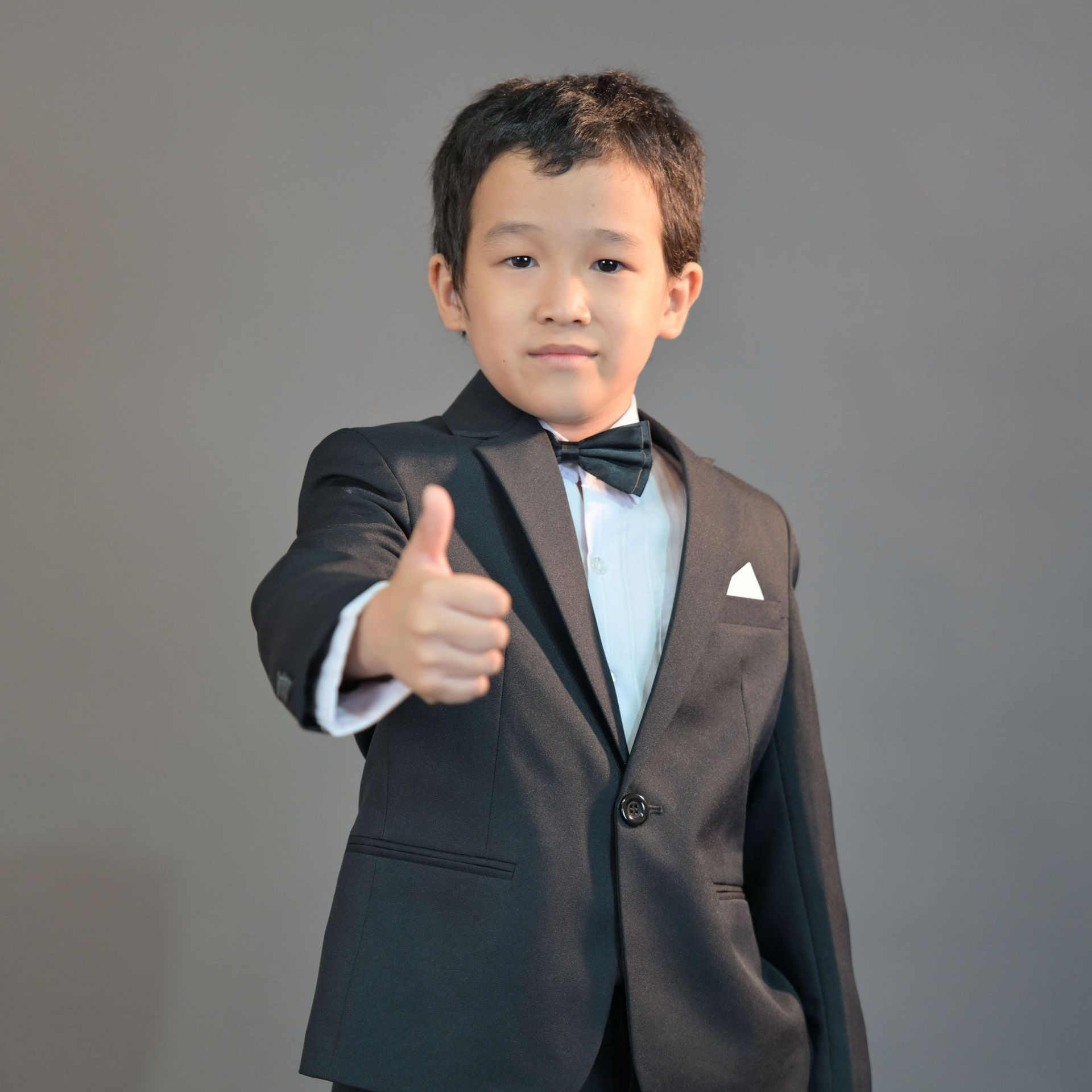 Child in a black suit giving a thumbs-up against a plain gray background