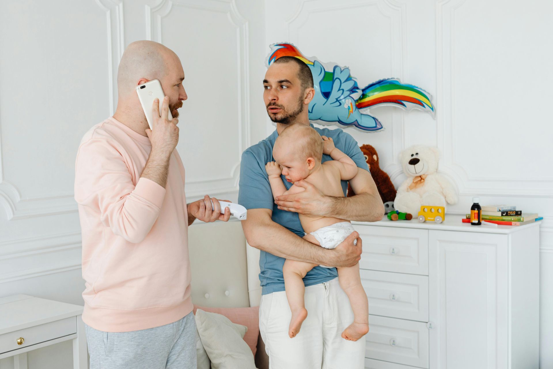 Two people in a bright nursery, one holding a baby, with a phone call and toys on a dresser.