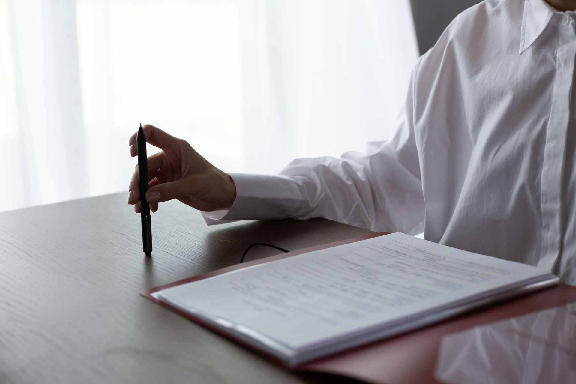 Hand holding a pen over an open notebook on a desk by a bright window