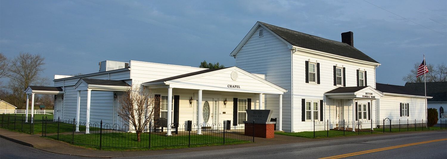A white, two-story house with a long, one-story attached side wing, seen from a street under a clear, pale blue sky.