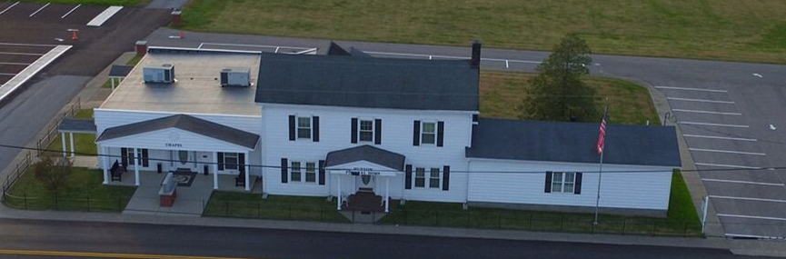 An aerial view of a white two-story building with a flat-roofed addition, a covered porch, and a parking lot.