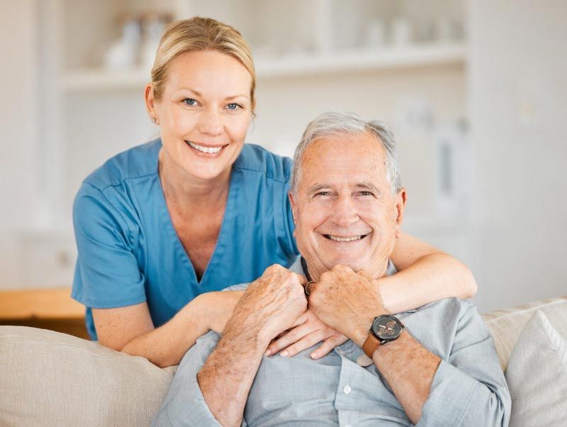 Caregiver in blue scrubs embraces a smiling elderly man sitting on a couch.