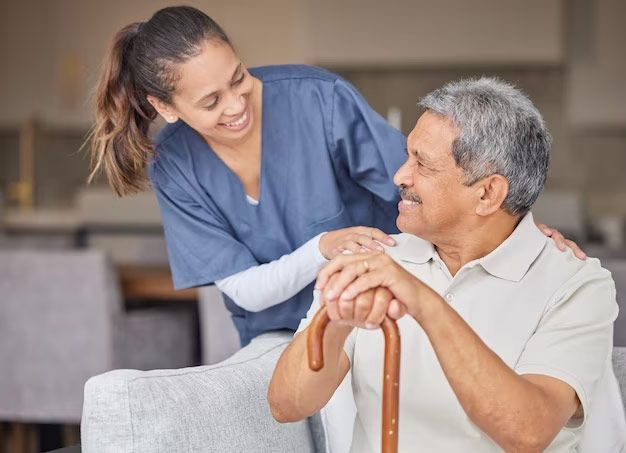 Woman in blue uniform smiles at an older man holding a cane. She places her hand on his shoulder. They are indoors.