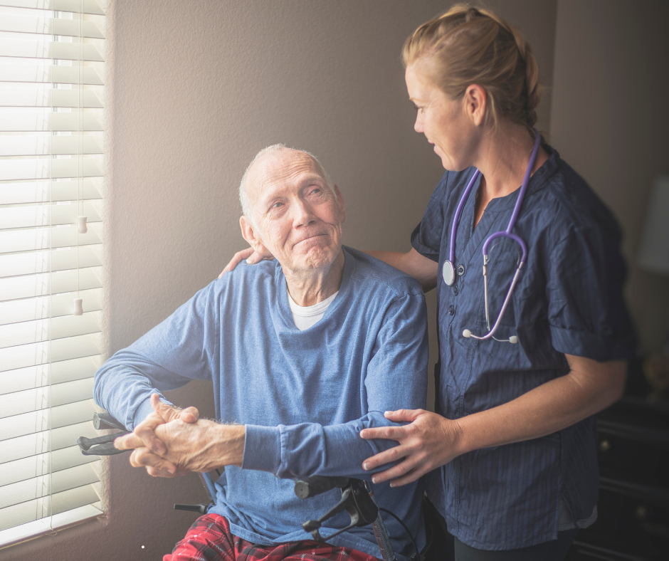 Woman in scrubs with stethoscope comforts elderly man in wheelchair, holding his hand.