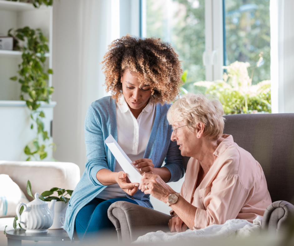 Woman assisting older person with paperwork, both looking at the document in a well-lit living room.