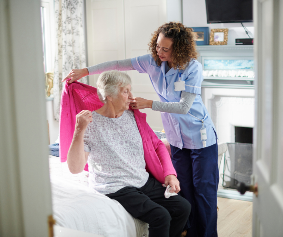 Caregiver helps an older person put on a pink jacket, seated on a bed.