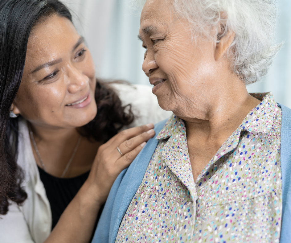 Woman with dark hair smiles, touches older woman's shoulder. Both women are inside, well-lit.