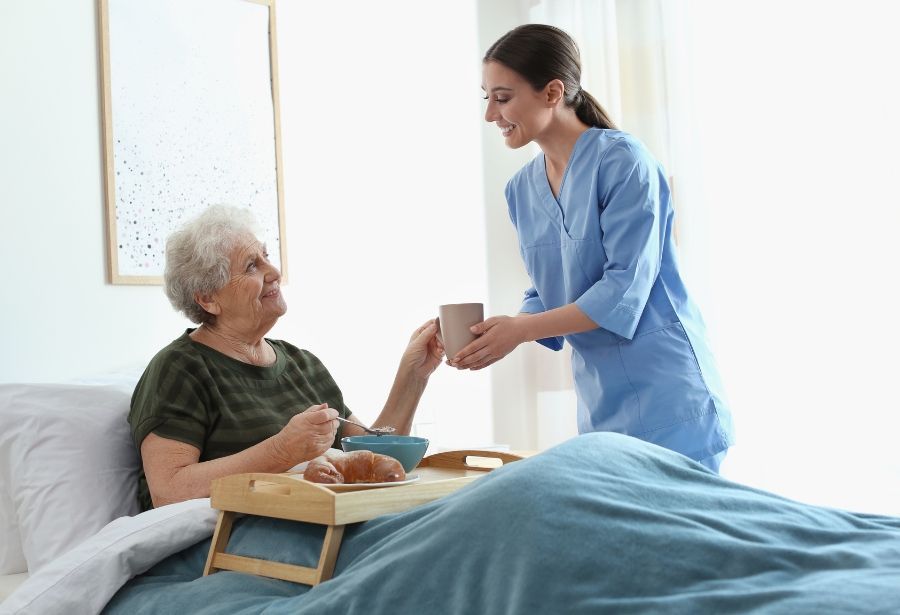 Caregiver serving a meal to a person in bed; a cup is being offered. Room setting.