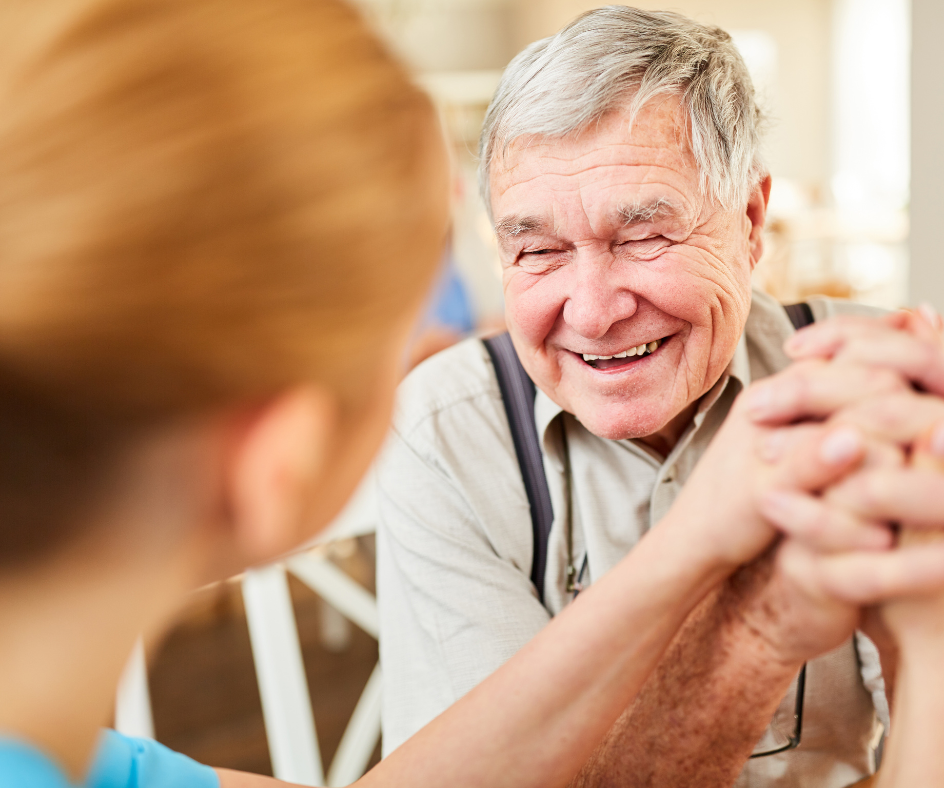 Elderly man smiles, holding hands with a person wearing a teal shirt. They are indoors.