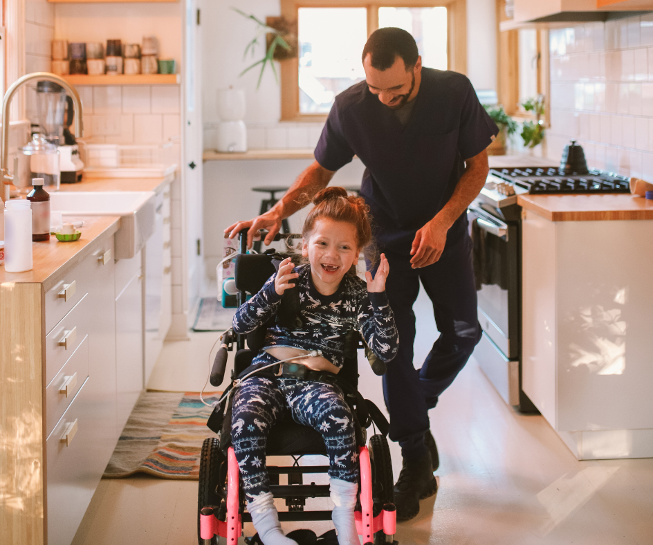 Man assists child in a wheelchair in a kitchen, both smiling. Bright setting with white cabinets and a window.