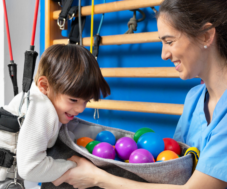 Boy in harness smiling, reaching for colorful balls from a basket held by smiling therapist in a therapy room.
