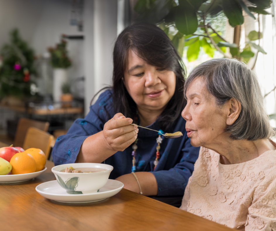Woman feeding an older person soup at a wooden table, in a home setting.