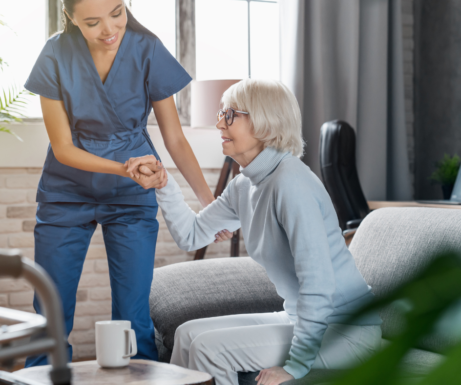 Caregiver assisting an older person to stand up from a sofa. Indoors, with a walking aid nearby.