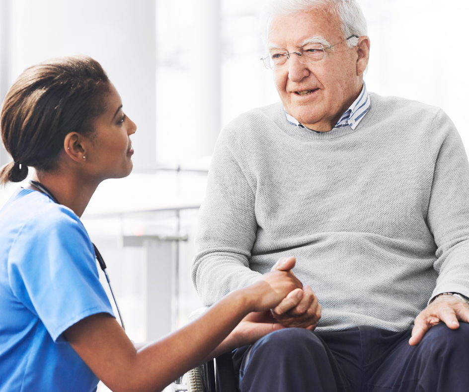 Nurse holding a patient's hand, speaking. Both are seated indoors, likely a medical setting.