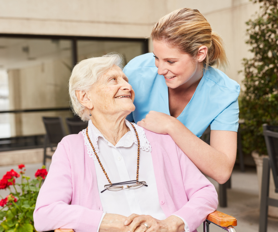 Woman in wheelchair smiles at a caregiver outdoors; the caregiver has her hand on the woman's shoulder.