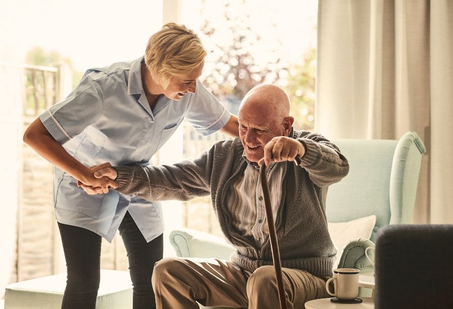 Caregiver assisting a person with a cane to stand up from a light blue chair in a sunlit room.