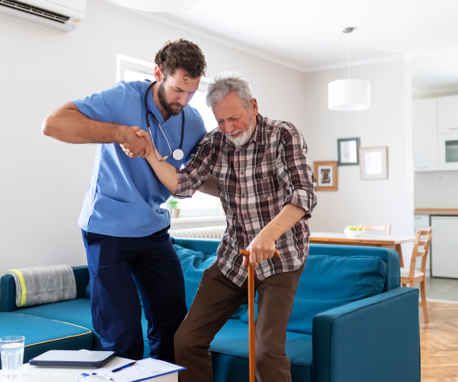 Caregiver assisting an older person with a cane to stand up; indoors, sofa in the background.