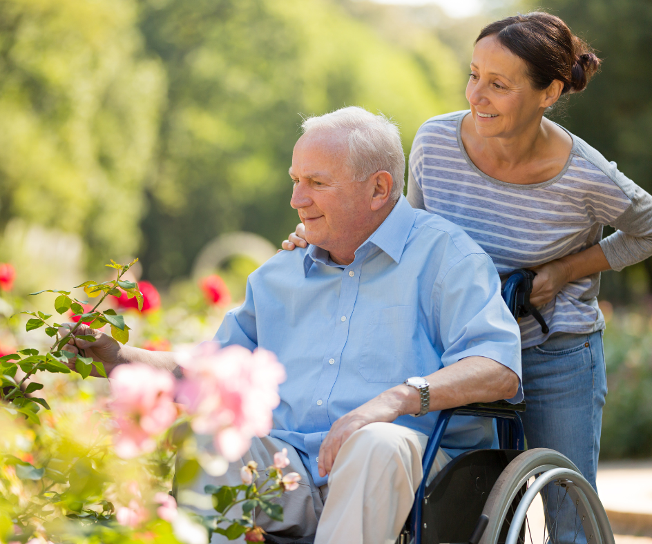 Man in wheelchair touching flowers, with woman standing beside him in a garden.