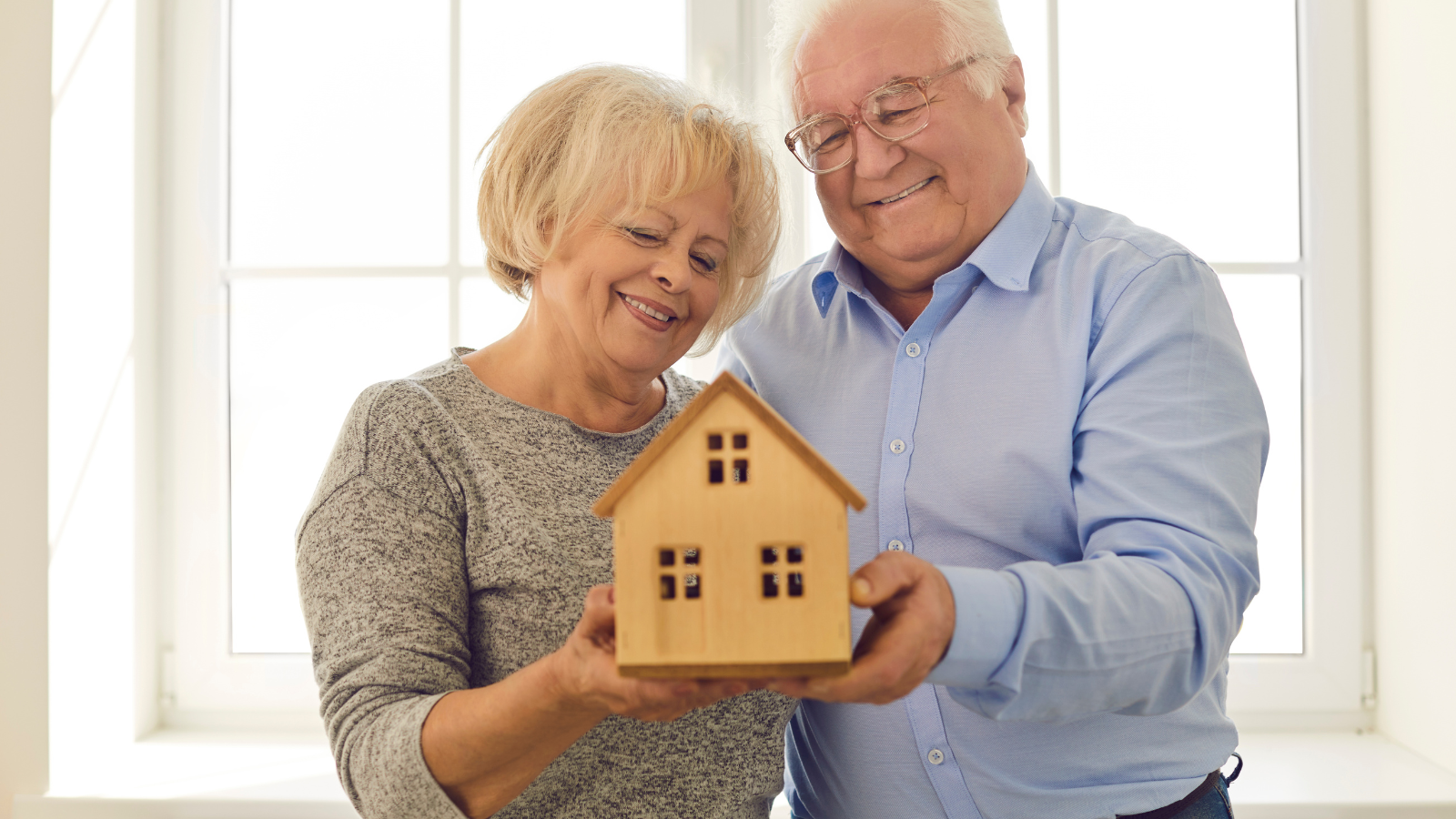 Elderly couple holding a small wooden house, smiling in front of a window.