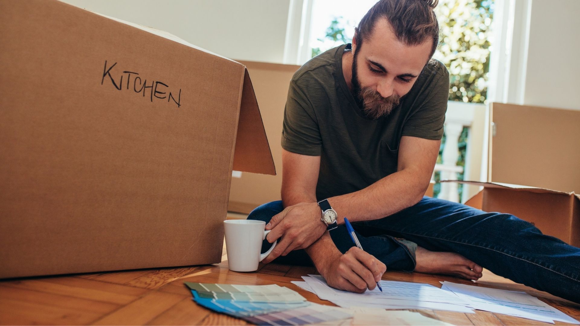 Man seated on floor, writing near moving boxes. A cup and paint swatches are next to him.