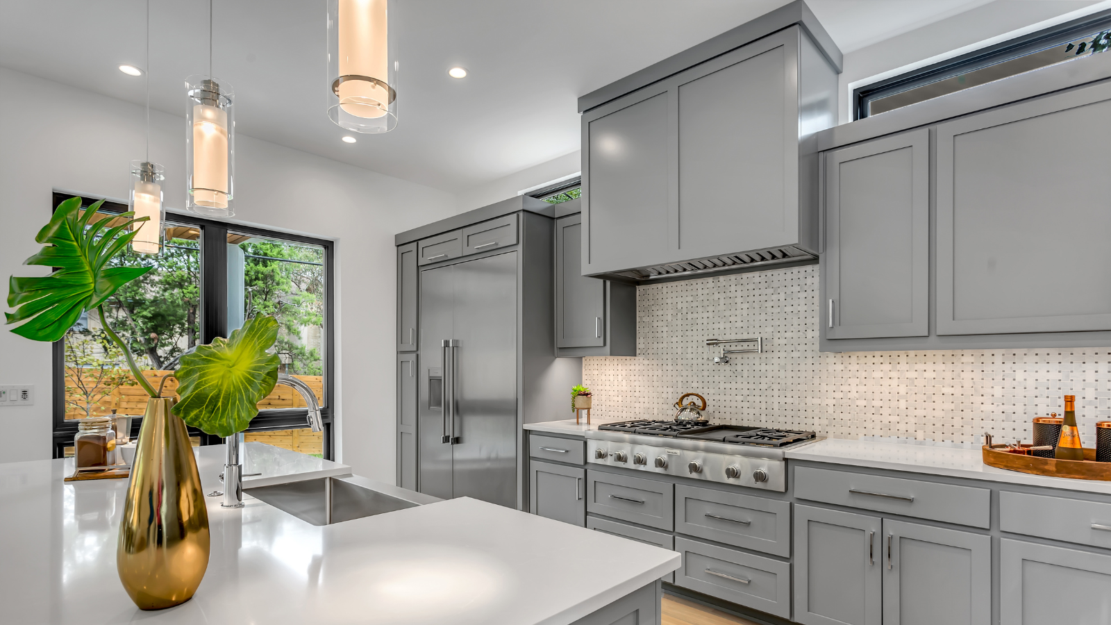 Modern gray kitchen with white countertops, stainless steel appliances, and large window overlooking greenery.