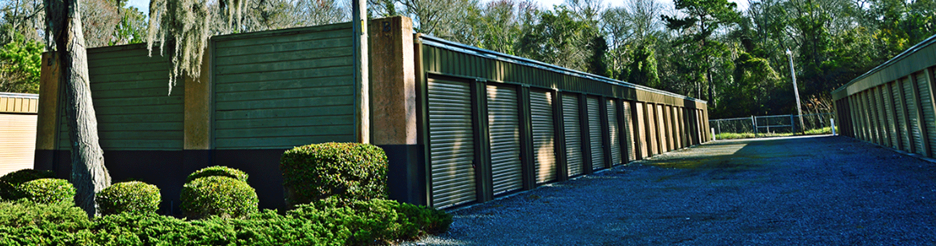 Long, modern, dark green storage units with slanted roofs. Gravel walkway and greenery in the foreground.