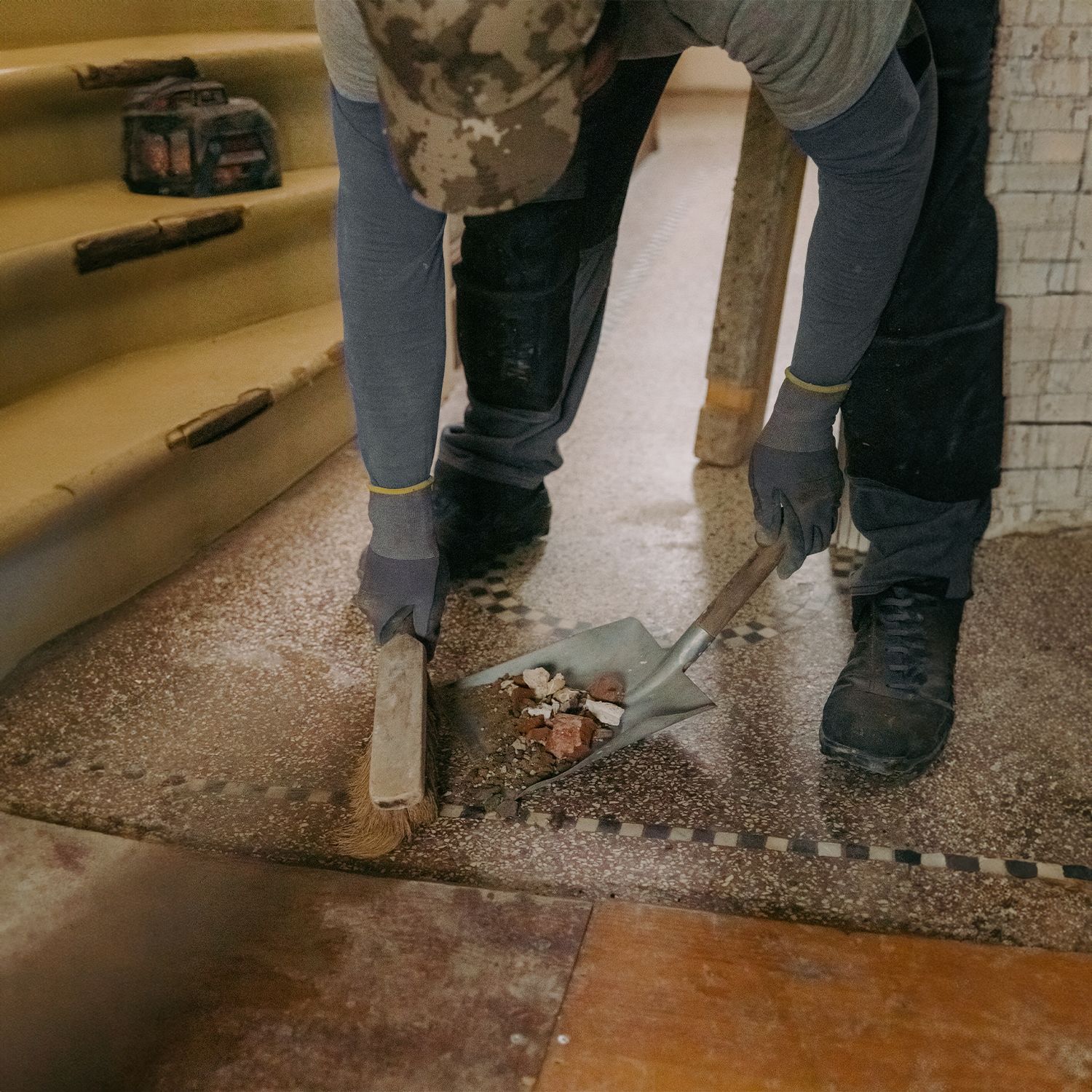 Persona raspando escombros del piso de baldosas con pala y cincel cerca de las escaleras.