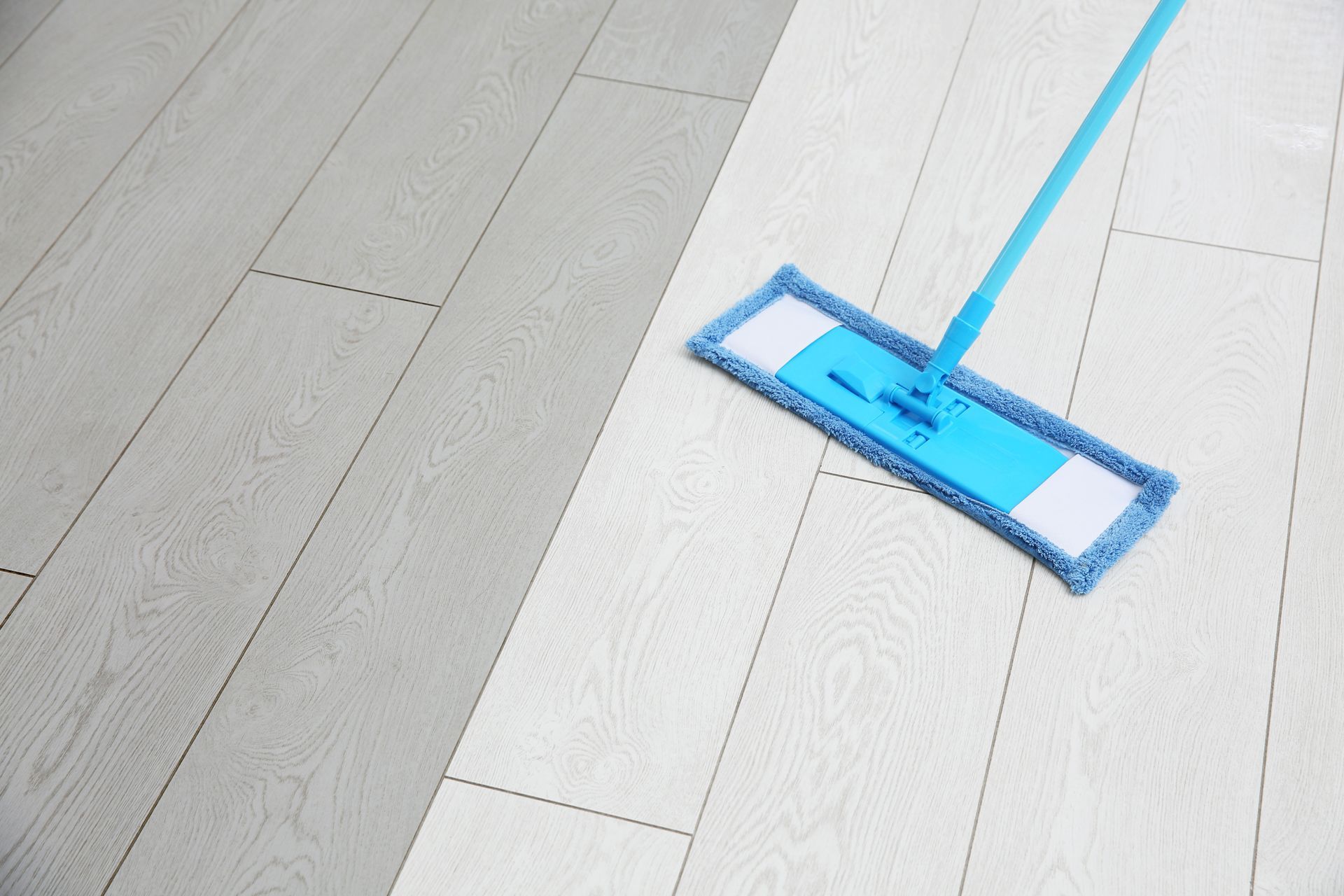 Una fregona azul limpia un suelo de madera clara. El suelo tiene una sección limpia y otra sin limpiar.
