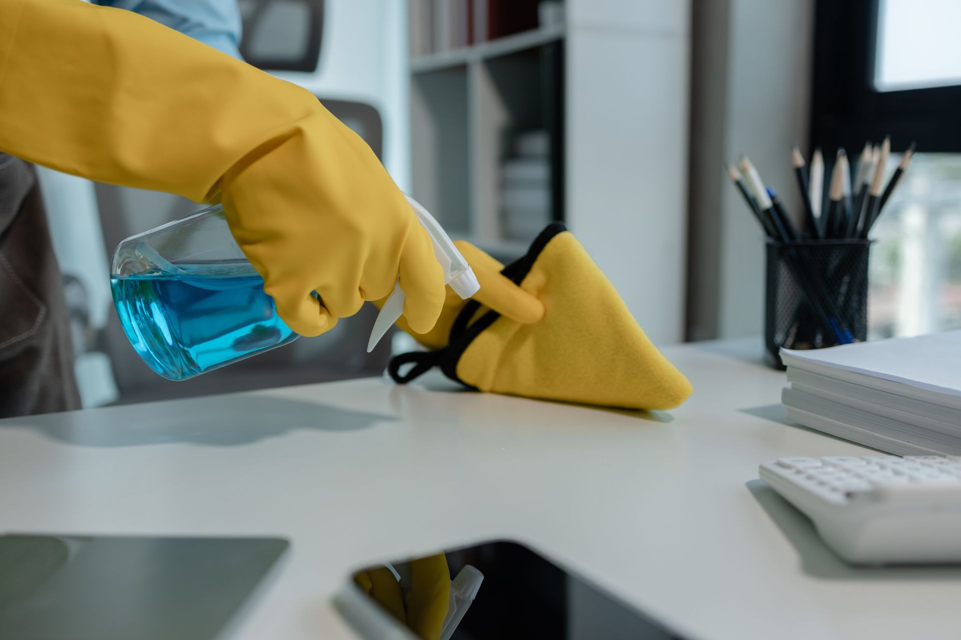Mano en guante amarillo rociando limpiador azul sobre un paño de limpieza amarillo, sobre un escritorio blanco.