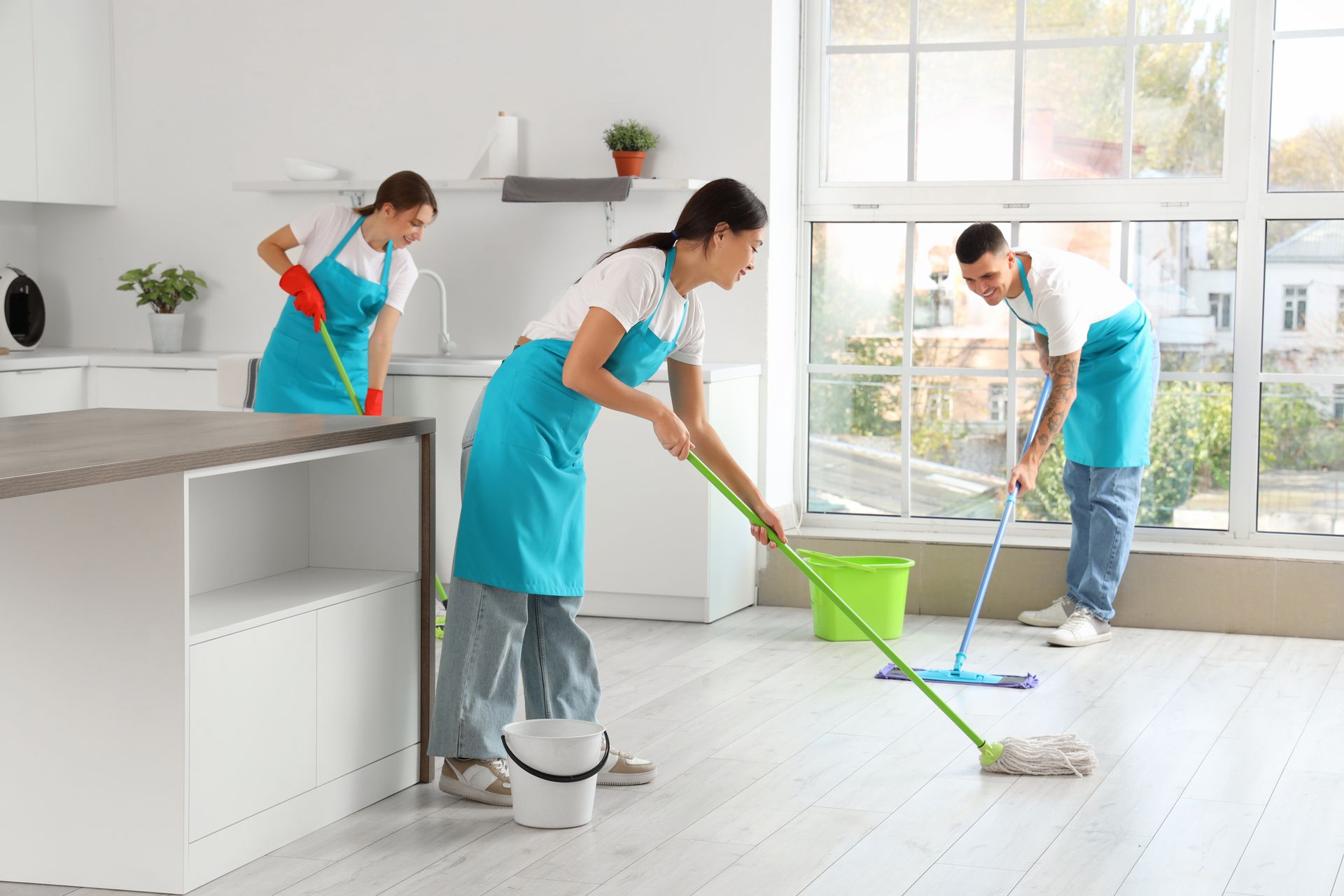 Tres personas limpiando una habitación bien iluminada: una trapeando, dos barriendo.
