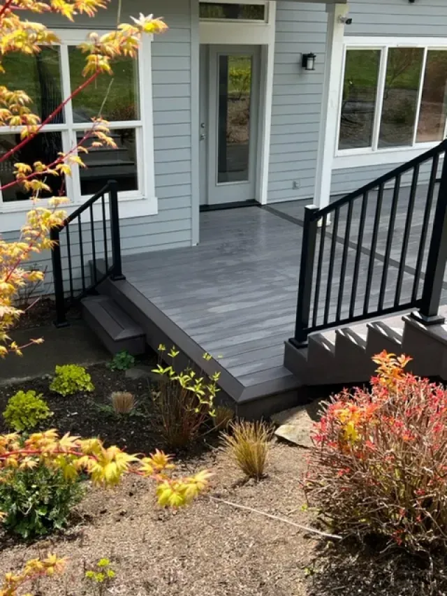 Gray composite deck with black railing and steps leading to a house entrance. Landscaping with plants in the foreground.