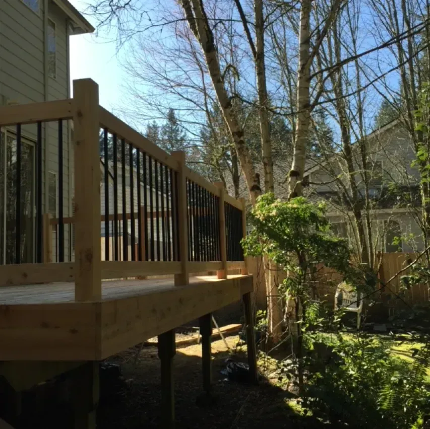 Wooden deck with black railing in a backyard with trees and a glimpse of a house.
