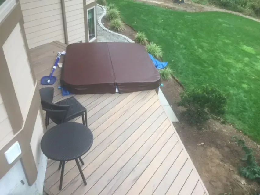 Hot tub with cover on a wooden deck next to green lawn, with a small table and chairs.