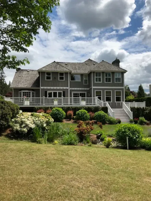 Two-story gray house with a wooden roof, white trim, and a grassy yard under a cloudy sky.