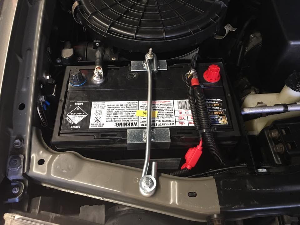 Car battery in a vehicle engine bay, with red and black terminals and a cooling fan above