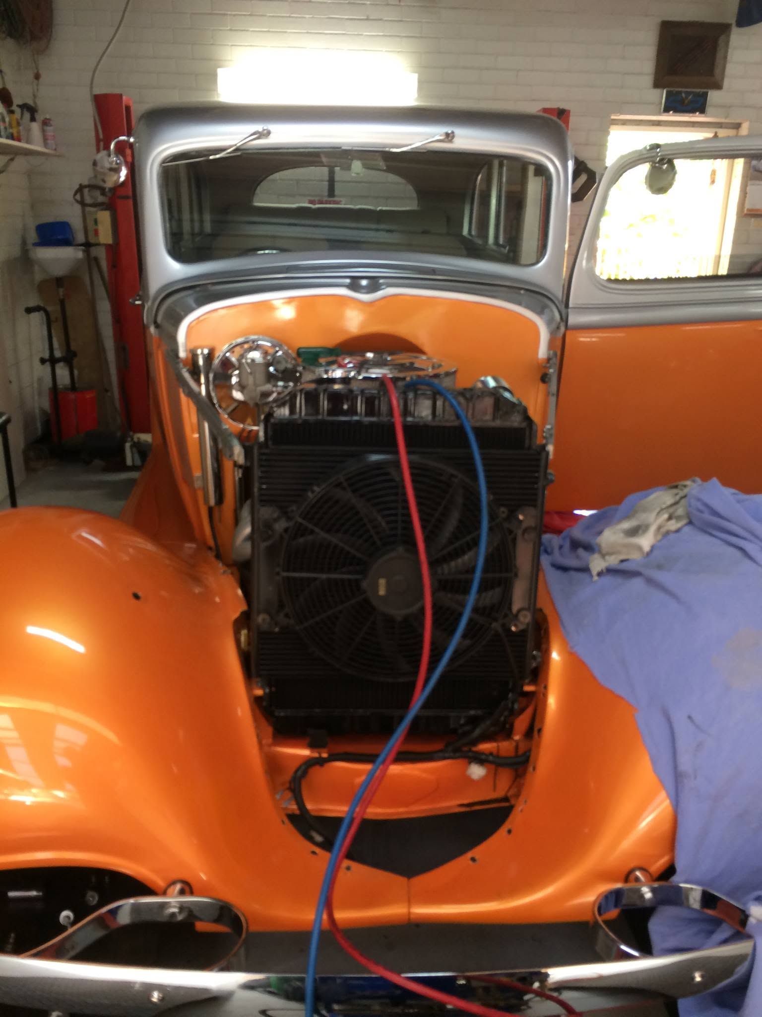 Orange vintage car in a garage, front grille exposed with jumper cables attached.
