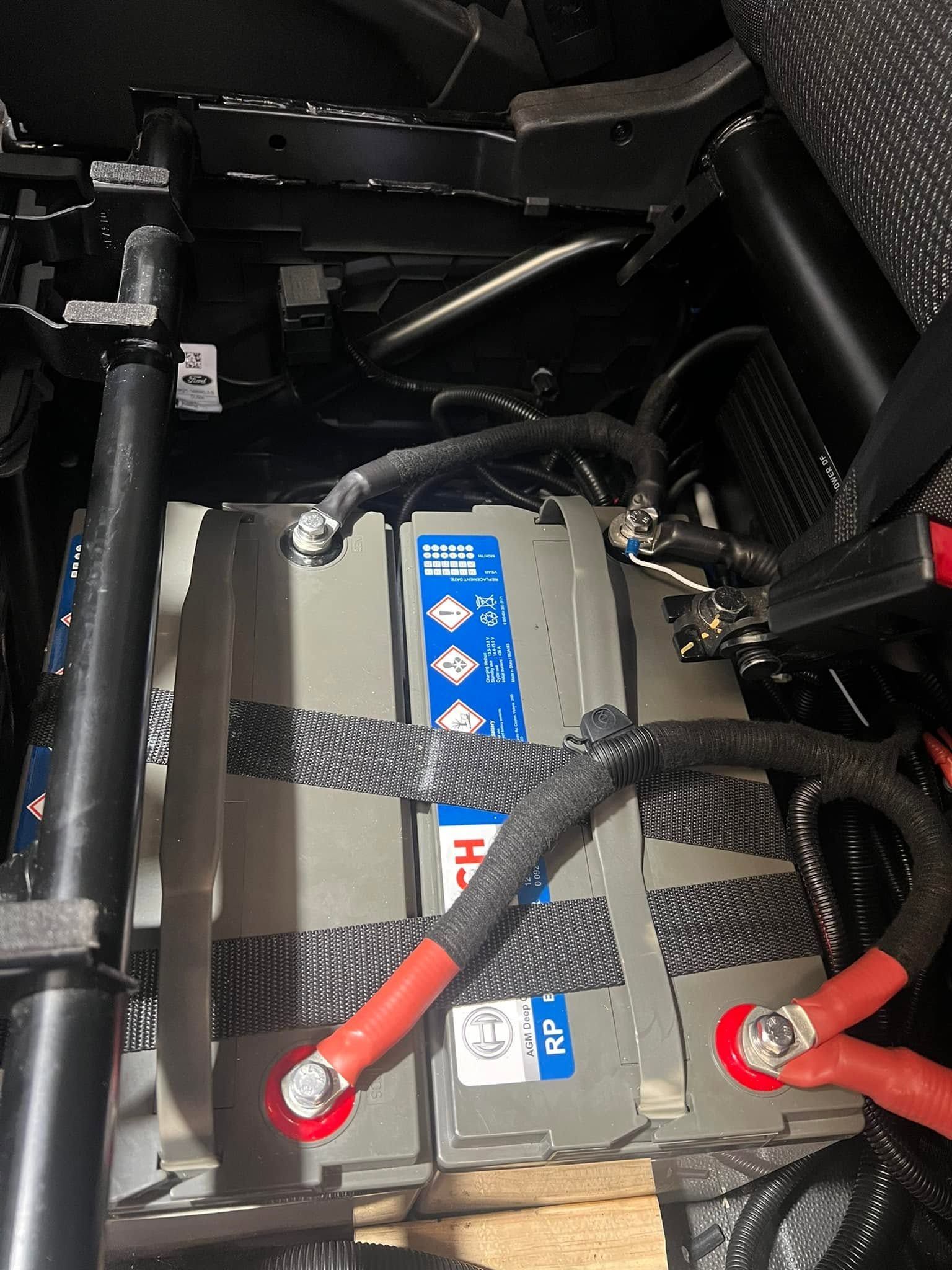 Battery compartment with black cables and red terminals secured by straps in a dark vehicle interior