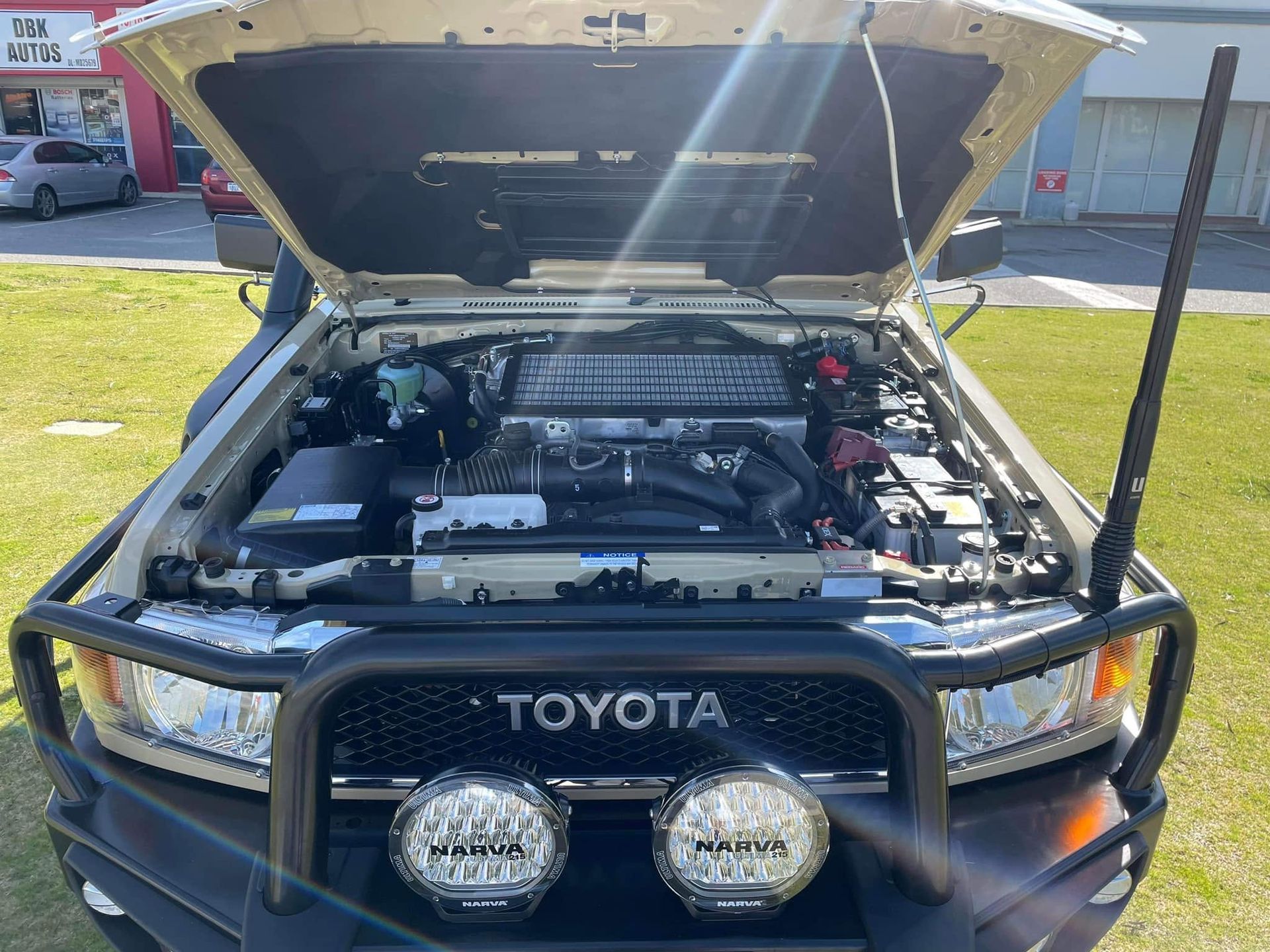 Front view of a Toyota pickup with hood open, showing engine bay and off-road lights in a driveway.