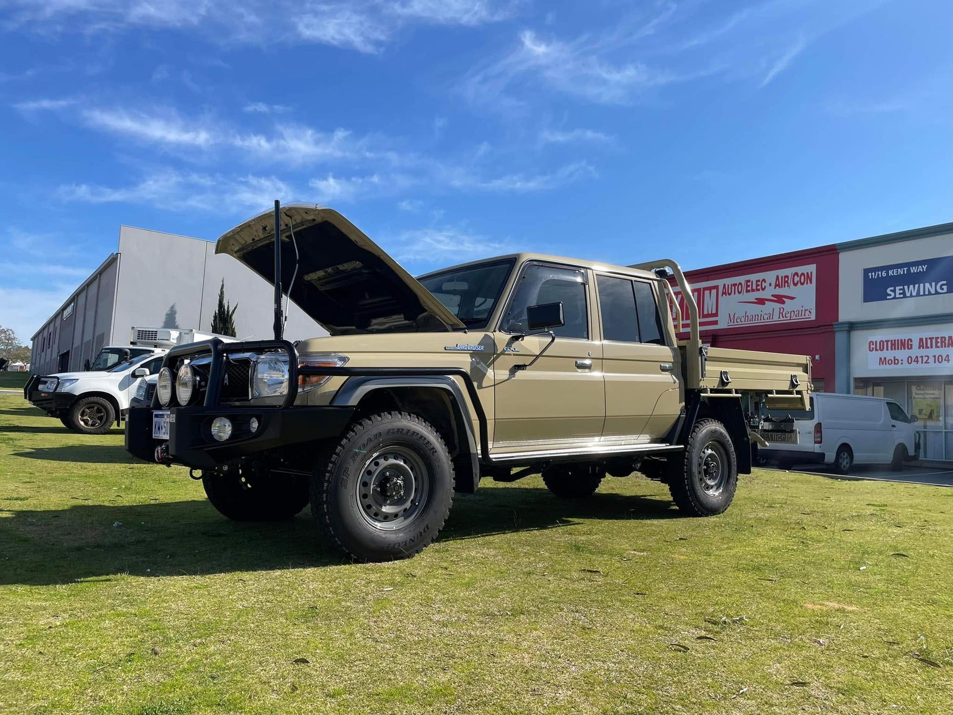 Tan pickup truck with open hood parked on grass at an outdoor event, with tents and signage behind it
