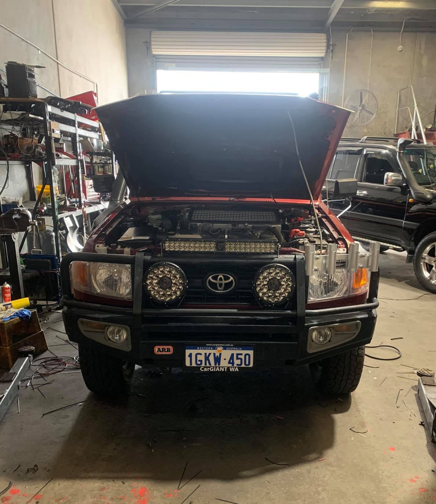 Black SUV with hood open in a garage, front view on a lift, with tools and another vehicle nearby