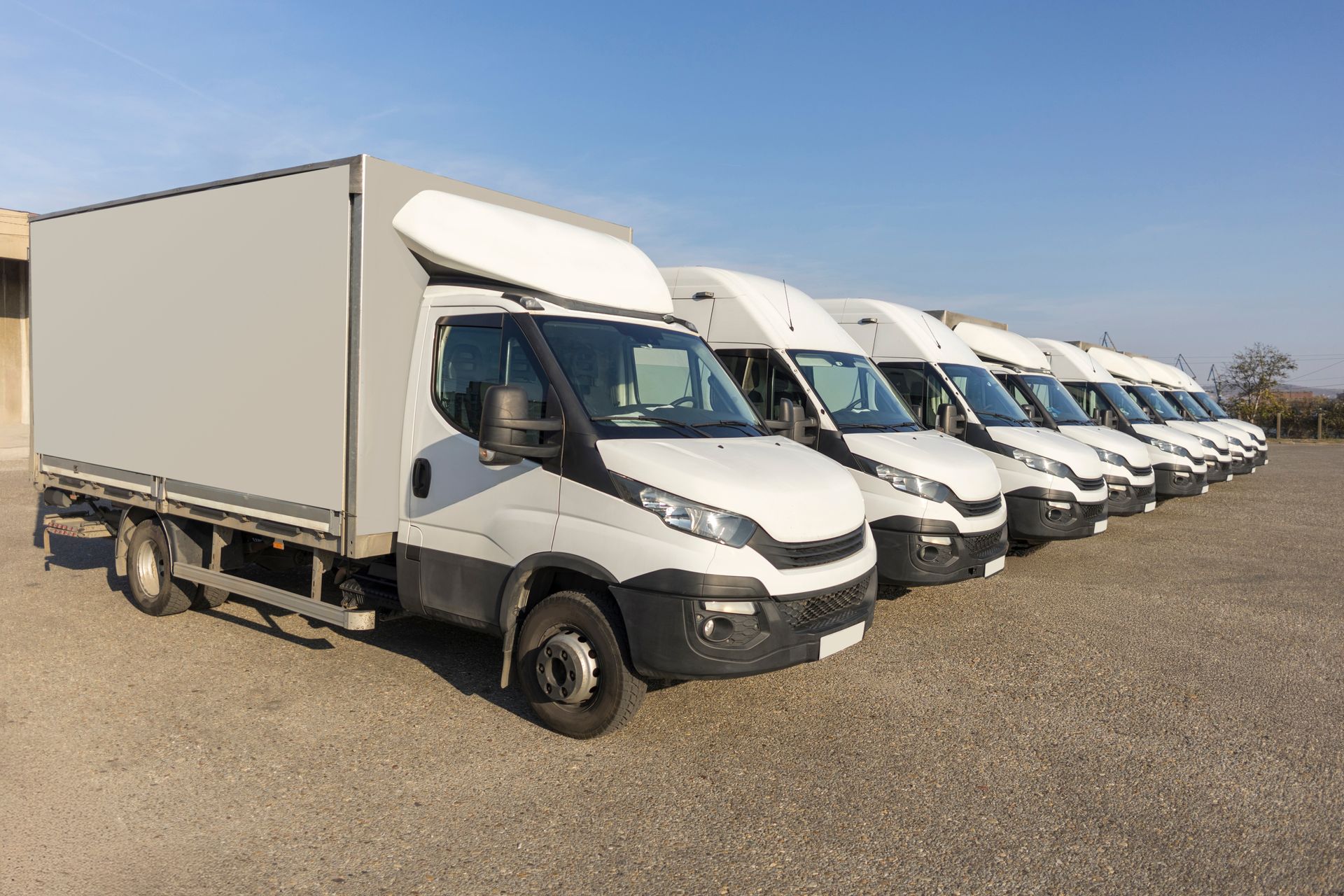 Row of white box trucks parked on a gravel lot under a clear blue sky