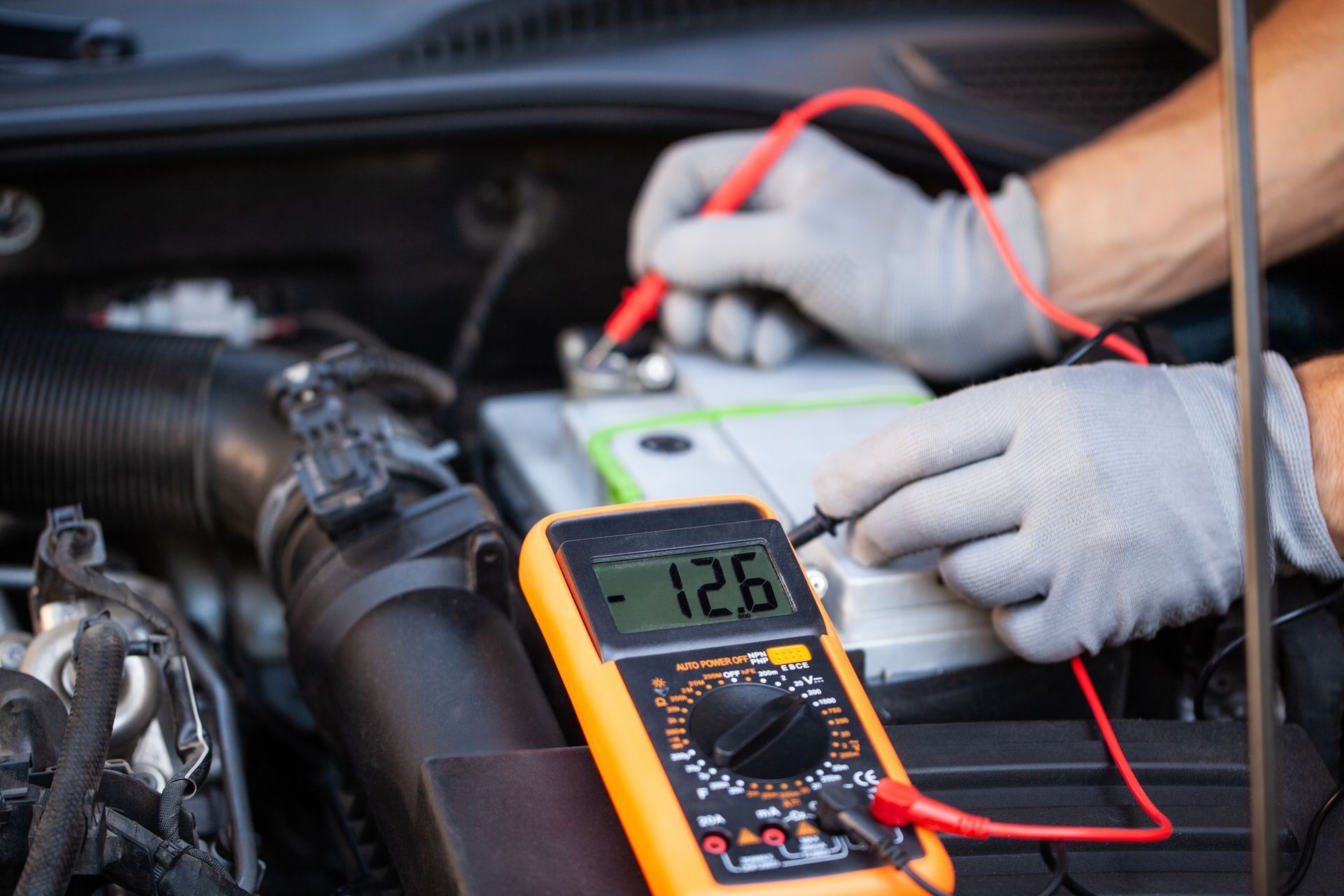 Mechanic testing a car battery with a digital multimeter and red probes in an engine bay Mechanic testing a car battery with a digital multimeter and red probes in an engine bay