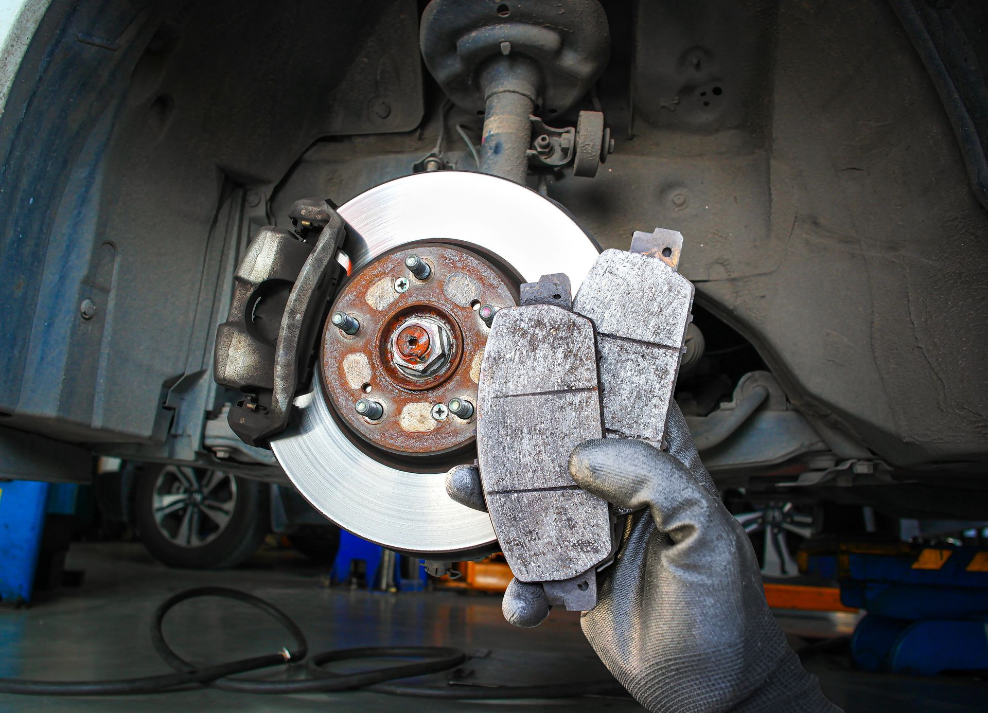 Brake rotor with worn brake pads held in a gloved hand under a car on a lift Brake rotor with worn brake pads held in a gloved hand under a car on a lift
