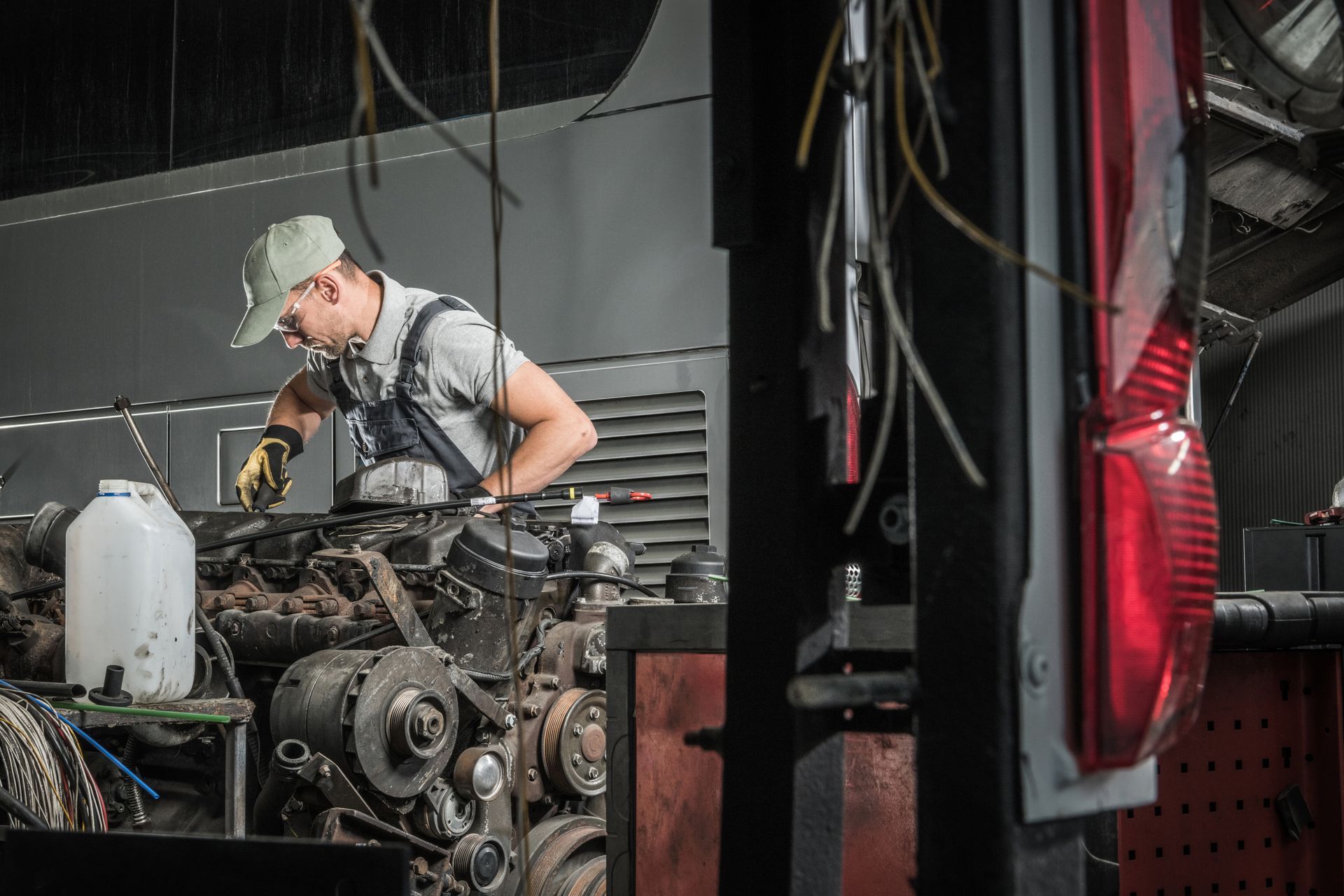 Mechanic in a cap works on an exposed engine in a dim garage, with tools and hanging cables nearby.