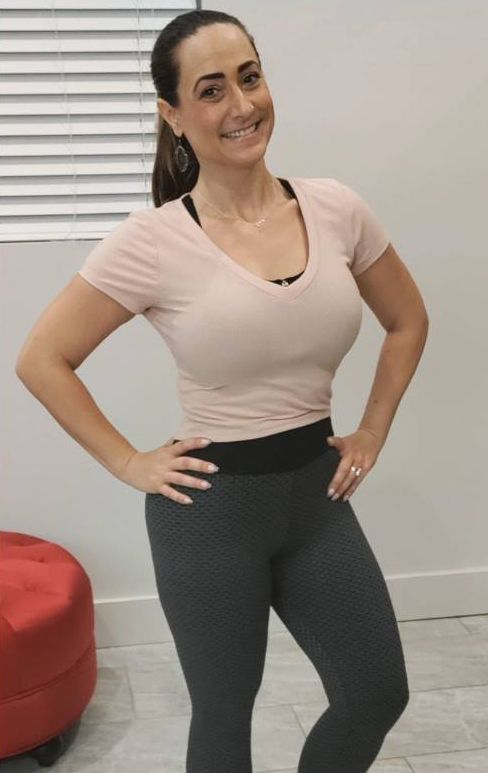 A woman in a pink shirt and grey leggings is standing with her hands on her hips.