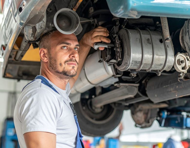 Mechanic working on a car from below, looking up. Wearing blue overalls and white shirt. Automotive shop.